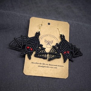 May include: A pair of black macrame bat earrings with red bead eyes and gold-toned hooks, displayed on a rectangular tan card featuring a butterfly logo and the text "Manifest the life you desire and deserve. almajewelry.etsy.com".