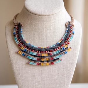 May include: A multi-layered statement necklace crafted from woven cord in shades of charcoal, tan, and light blue. The design features rows of small, colorful glass beads in red, yellow, and turquoise, arranged in a wide, curved, collar-like shape.