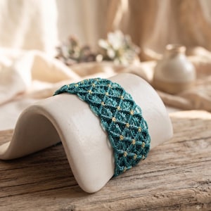 May include: A wide bracelet featuring a teal macrame cord design with small, integrated gold-colored beads arranged in a repeating diamond pattern. The woven textile accessory displays a structured, knotted texture throughout its entire surface.