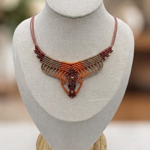 May include: A macrame necklace featuring a geometric pattern in shades of dark brown, tan, and orange. The design includes intricate knotted cord work, thin cord straps, and small metallic gold-toned beads integrated into the structure.