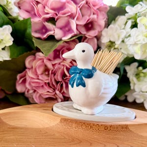 May include: A white ceramic duck toothpick holder with a blue bow, holding wooden toothpicks. The duck sits on a small, white, oval-shaped dish. The background features pink and white flowers and a wooden tray.
