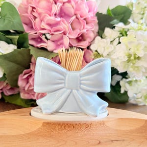 May include: A light blue ceramic toothpick holder shaped like a bow, filled with wooden toothpicks. The holder sits on a white base, with pink and white flowers in the background. The holder is a decorative item.