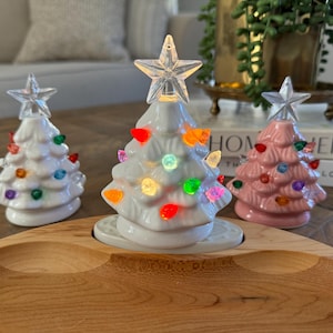 May include: Three ceramic Christmas trees with colorful lights. The trees are white, pink, and white. The trees are sitting on a wooden tray.
