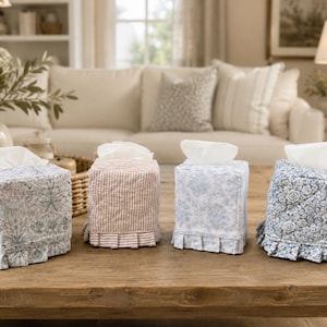 May include: Four square fabric tissue box covers with white tissues protruding from the top. Each cover features a quilted texture and a pleated ruffle along the bottom edge. The designs include floral patterns in blue and white and a pink striped pattern.