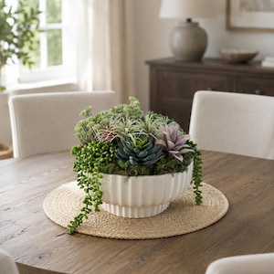 May include: A white ceramic bowl with a vertical ribbed texture containing an arrangement of various artificial succulents and greenery. The plants include trailing green vines, dark green rosettes, and light purple-tipped succulent leaves.