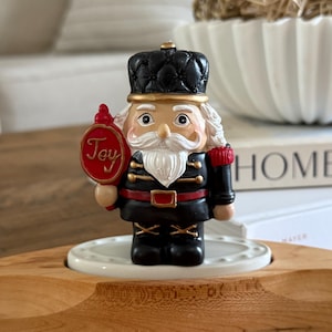 Puede incluir: Figura decorativa de cascanueces con uniforme clásico, sombrero y abrigo negros, detalles rojos y barba blanca. La figura sostiene un letrero rojo con la palabra "Joy".