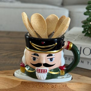 Puede incluir: Una taza de cerámica con forma de cascanueces con un uniforme verde y dorado, un sombrero negro y una cara blanca. La taza contiene un juego de cucharas de madera.