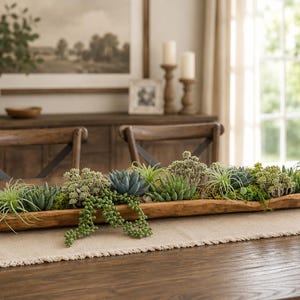 May include: A long, narrow, natural wood dough bowl filled with an assortment of artificial succulents and greenery. The arrangement features various shades of green, including spiky air plants, rounded rosettes, and trailing beaded stems.