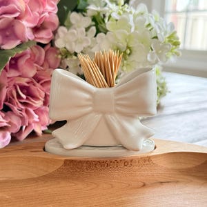 Può includere: Un portastuzzicadenti in ceramica bianca a forma di fiocco, riempito di stuzzicadenti in legno. Il supporto è posto su un vassoio di legno, con fiori rosa e bianchi sullo sfondo. Un oggetto decorativo per la cucina.