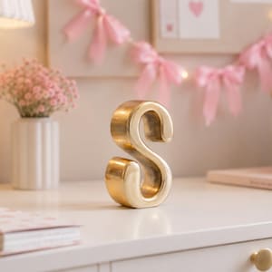 May include: A freestanding, three-dimensional letter S with a smooth, polished gold-tone metallic finish. The letter features a rounded, cursive-inspired typeface with a consistent thickness throughout its structure, standing upright on a flat surface.