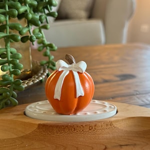 May include: A small, bright orange pumpkin figurine featuring a smooth, glossy finish and a brown stem. The pumpkin is adorned with a white ribbon tied in a bow at the top and is mounted on a small, oval, white ceramic base with a beaded edge.