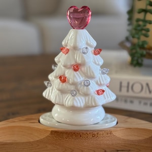 May include: A white ceramic Christmas tree with pink and clear lights and a pink heart on top. The tree is sitting on a wooden base.
