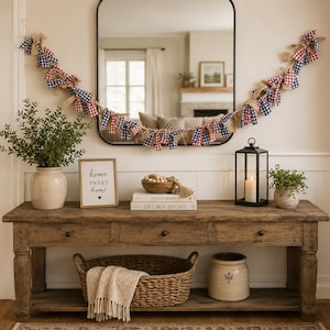 May include: A fabric rag garland featuring red, white, and blue gingham patterns mixed with natural burlap strips, draped across a rectangular mirror above a wooden console table with three drawers, a woven basket, and decorative home accents.