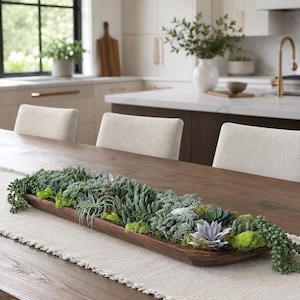 May include: A long, narrow, dark brown wooden dough bowl filled with a variety of green succulents, air plants, and bright green moss. The arrangement features diverse textures and shapes, with trailing plants hanging over the ends of the bowl.