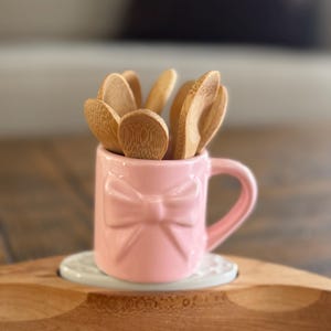 Może przedstawiać: Różowy ceramiczny kubek z motywem kokardy, wypełniony małymi drewnianymi łyżkami. Kubek stoi na białej podstawce i jest umieszczony na drewnianej tacy. Łyżki są wykonane z jasnego drewna.