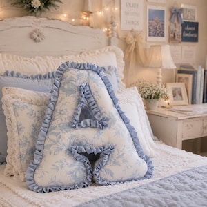 May include: A letter A shaped throw pillow featuring a white fabric with a light blue floral toile pattern. The pillow is edged with a ruffled light blue fabric trim that follows the entire contour of the letter shape, creating a decorative border.