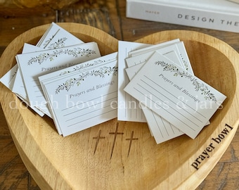 May include: A wooden heart-shaped bowl with the words "prayer bowl" engraved on the side. The bowl is filled with small white cards with the words "Prayers and Blessings" printed on them. Three crosses are carved into the bottom of the bowl.