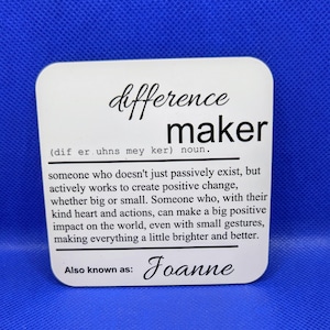 May include: A white square coaster with rounded corners. The coaster features the words "difference maker" in a stylish font, along with a definition. The name "Joanne" is also printed on the coaster.