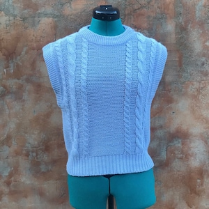 Peut inclure: Un gilet en tricot de couleur bleu clair avec un col rond. Le gilet est présenté sur un mannequin vert avec un support noir, sur un fond texturé marron et orange. Le gilet présente un panneau central et des détails en tricot torsadé.