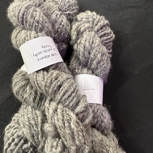 Puede incluir: Dos madejas de hilo gris grueso, etiquetadas como "100% Alpaca Trudy 50 yards". El hilo tiene un aspecto grueso y texturizado, ideal para proyectos de tejido o ganchillo. El hilo es de un color gris suave y neutro.