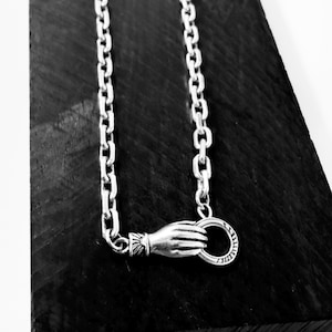 May include: A silver chain necklace featuring a clasp designed as a hand holding a circular ring. The hand motif includes detailed wrist cuff engravings, and the chain consists of uniform oval links, creating a cohesive metallic accessory.