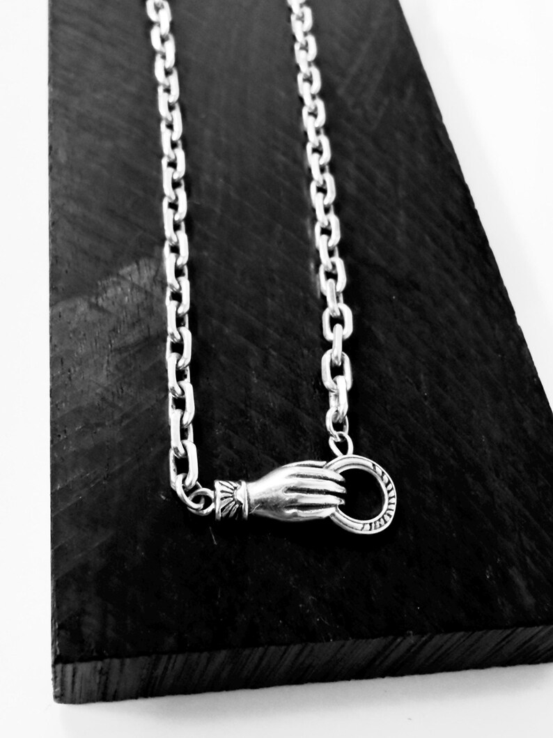 May include: A silver chain necklace featuring a clasp designed as a hand holding a circular ring. The hand motif includes detailed wrist cuff engravings, and the chain consists of uniform oval links, creating a cohesive metallic accessory.