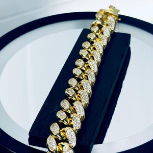 May include: A gold-toned cuban link bracelet featuring a series of interlocking oval links. Each link is encrusted with numerous small, clear, reflective stones that cover the top surface. The bracelet includes a rectangular box clasp closure.