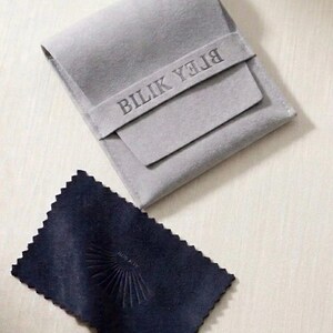 May include: A light gray felt pouch with a flap closure featuring the embossed text BILIK YEJIA, accompanied by a rectangular dark navy blue polishing cloth with pinked edges and a small embossed sunburst design in the center.