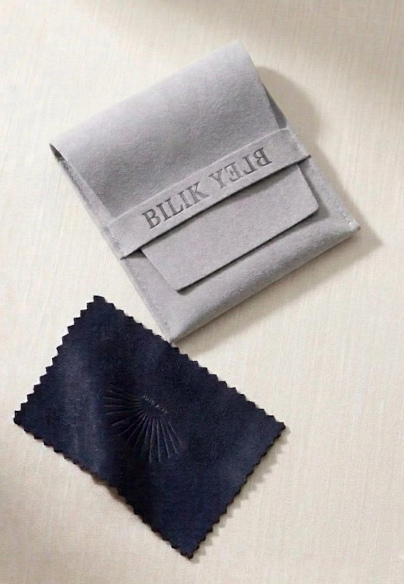 May include: A light gray felt pouch with a flap closure featuring the embossed text BILIK YEJIA, accompanied by a rectangular dark navy blue polishing cloth with pinked edges and a small embossed sunburst design in the center.