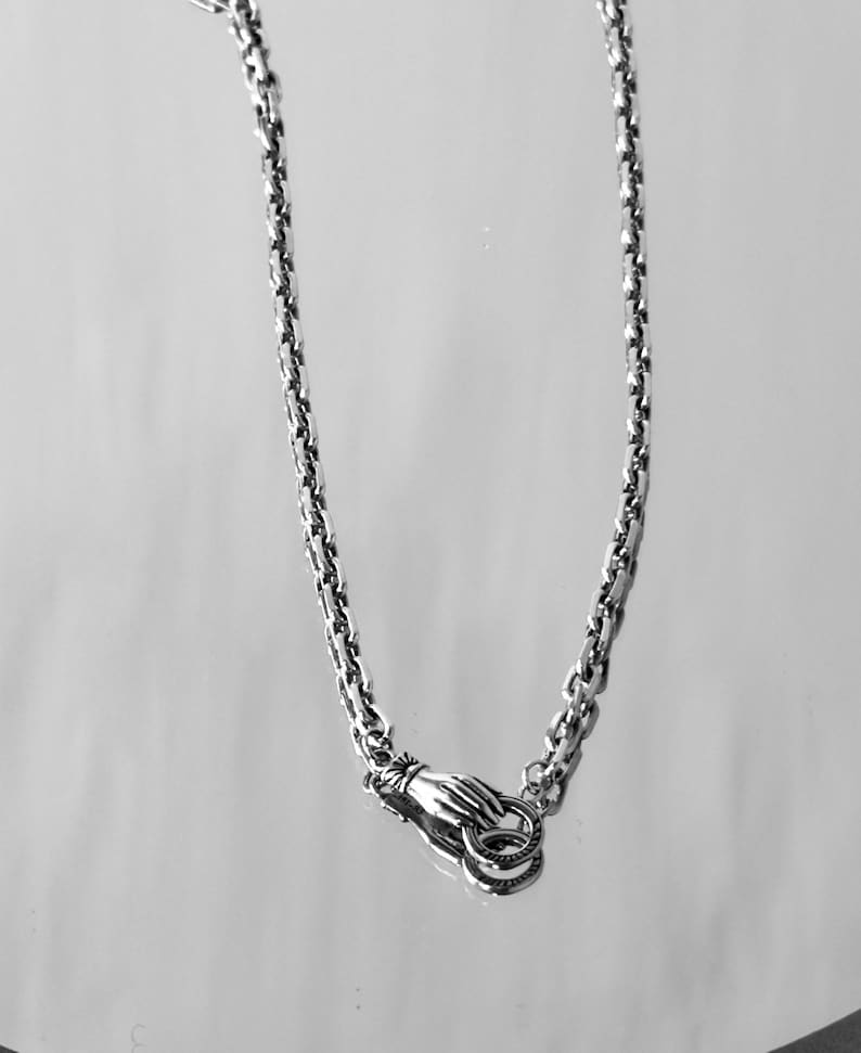 May include: A silver-toned metal chain necklace featuring a clasp designed as a hand holding two small circular rings. The chain consists of interlocking oval links with a polished finish, creating a continuous loop that hangs downward.