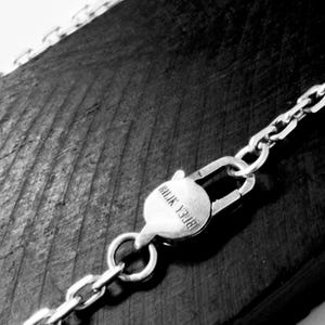 May include: A silver-toned metal chain necklace featuring a circular lobster claw clasp. The clasp is engraved with the text "BILIK YJ08". The chain consists of rectangular interlocking links that extend across the frame in a diagonal orientation.