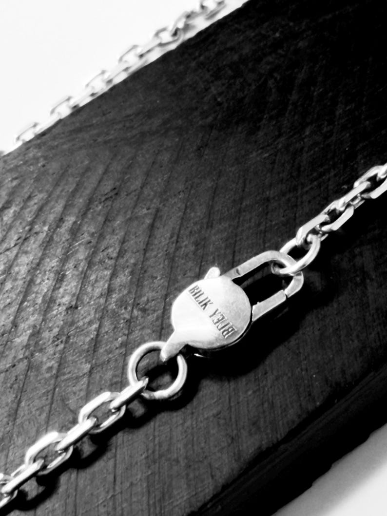 May include: A silver-toned metal chain necklace featuring a circular lobster claw clasp. The clasp is engraved with the text "BILIK YJ08". The chain consists of rectangular interlocking links that extend across the frame in a diagonal orientation.