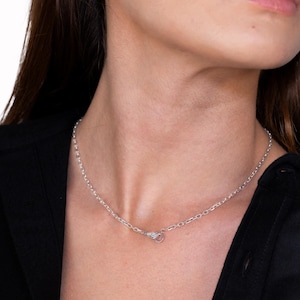 May include: A silver-toned chain necklace featuring a unique clasp designed in the shape of a small hand. The metallic links are uniform and circular, creating a simple yet distinct look that rests around the neck area with a polished finish.