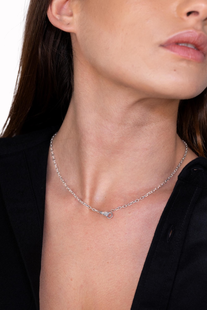 May include: A silver-toned chain necklace featuring a unique clasp designed in the shape of a small hand. The metallic links are uniform and circular, creating a simple yet distinct look that rests around the neck area with a polished finish.