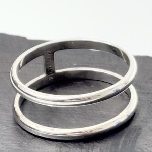 K&ouml;nnte beinhalten: Ein polierter silberfarbener Ring mit einem Doppelband-Design. Die beiden kreisf&ouml;rmigen B&auml;nder sind an einer Seite durch einen einzelnen vertikalen Steg verbunden, wodurch ein offener Raum zwischen den B&auml;ndern entsteht. Die Metalloberfl&auml;che ist glatt.