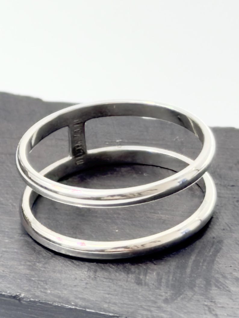 K&ouml;nnte beinhalten: Ein polierter silberfarbener Ring mit einem Doppelband-Design. Die beiden kreisf&ouml;rmigen B&auml;nder sind an einer Seite durch einen einzelnen vertikalen Steg verbunden, wodurch ein offener Raum zwischen den B&auml;ndern entsteht. Die Metalloberfl&auml;che ist glatt.