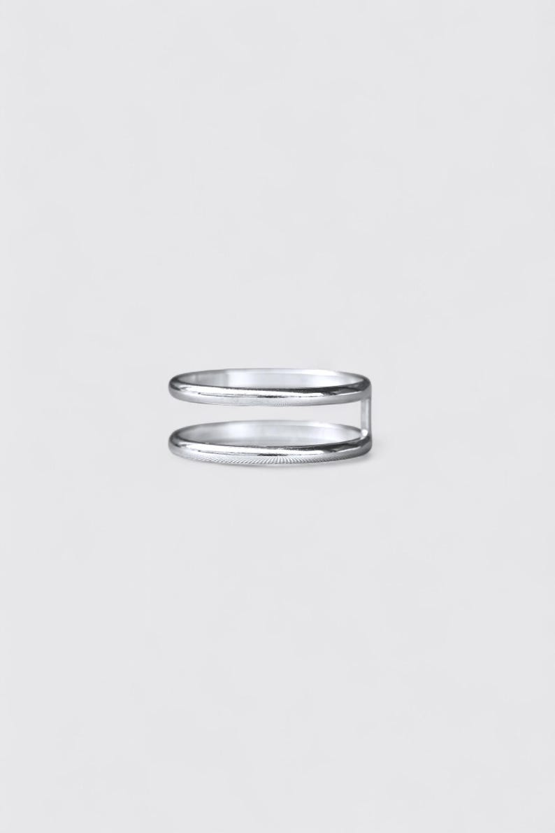 K&ouml;nnte beinhalten: Ein silberfarbener Ring mit einem Doppelband-Design. Die zwei d&uuml;nnen, glatten, kreisf&ouml;rmigen B&auml;nder sind an einer Seite durch einen einzelnen vertikalen Steg verbunden, was eine offene, parallele Struktur mit polierter, gl&auml;nzender Oberfl&auml;che ergibt.