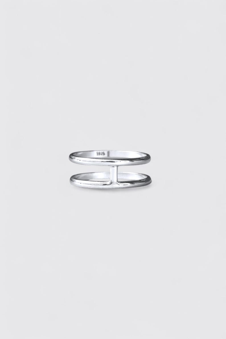 K&ouml;nnte beinhalten: Ein Ring aus poliertem Silber mit zwei parallelen B&auml;ndern, die durch einen vertikalen Steg verbunden sind. Auf der Innenseite des oberen Bandes ist deutlich die Pr&auml;gung 925 zu sehen, die auf das Sterlingsilber dieses schlichten Schmuckst&uuml;cks hinweist.