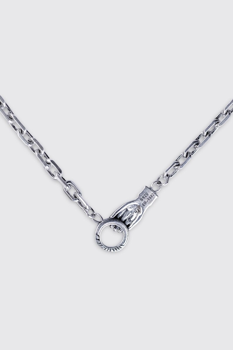 May include: A silver-toned metal chain necklace featuring a unique clasp shaped like a hand holding a circular ring. The clasp is engraved with the text S925 and ER-1882. The chain consists of rectangular links with a polished finish.