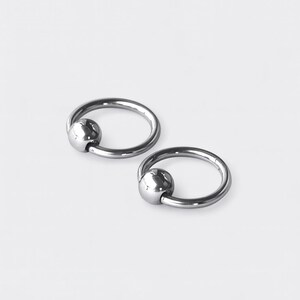May include: Two silver-colored captive bead rings, each consisting of a circular hoop with a single polished spherical bead held in place by the tension of the ring. The metal has a smooth, reflective finish and a metallic gray appearance.