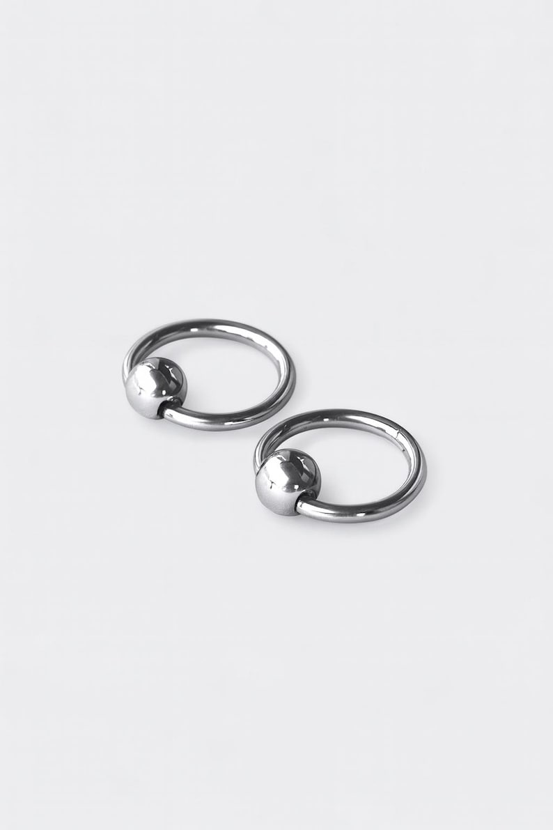 May include: Two silver-colored captive bead rings, each consisting of a circular hoop with a single polished spherical bead held in place by the tension of the ring. The metal has a smooth, reflective finish and a metallic gray appearance.