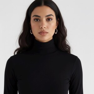 May include: A black long-sleeve shirt featuring a high turtleneck collar. The garment is made of a solid black fabric with a smooth texture, providing full coverage up to the neck. The design is simple, focusing on the fitted silhouette and collar.