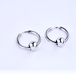 May include: A pair of silver-toned hoop earrings, each featuring a single polished spherical bead threaded onto the circular wire. The earrings have a smooth, reflective metallic finish and a hinged closure mechanism for secure wear.
