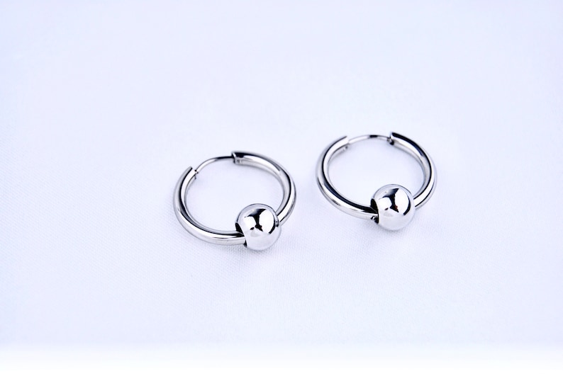 May include: A pair of silver-toned hoop earrings, each featuring a single polished spherical bead threaded onto the circular wire. The earrings have a smooth, reflective metallic finish and a hinged closure mechanism for secure wear.