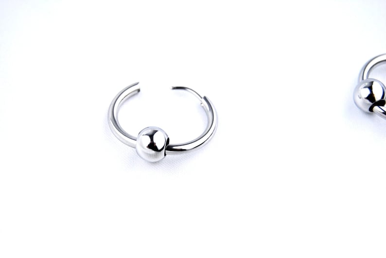 May include: A pair of silver-toned hoop earrings, each featuring a single polished, spherical bead threaded onto the circular wire band. The earrings have a smooth, reflective metallic finish and a simple, open-ended design for easy wear.