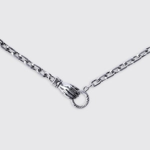 May include: A silver-toned metal chain necklace featuring a unique clasp mechanism designed as a hand holding a circular ring. The chain consists of rectangular links with a polished finish, and the hand clasp detail is textured.