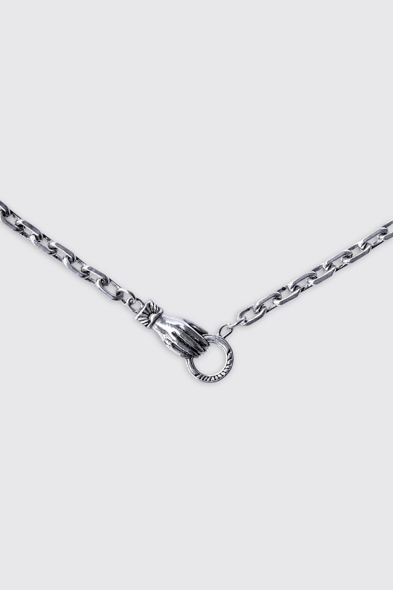 May include: A silver-toned metal chain necklace featuring a unique clasp mechanism designed as a hand holding a circular ring. The chain consists of rectangular links with a polished finish, and the hand clasp detail is textured.