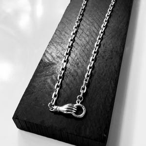 May include: A silver chain necklace featuring a clasp designed as a hand holding a circular ring. The metallic links are uniform and connected to the hand-shaped fastener, which secures the necklace with a circular loop mechanism.