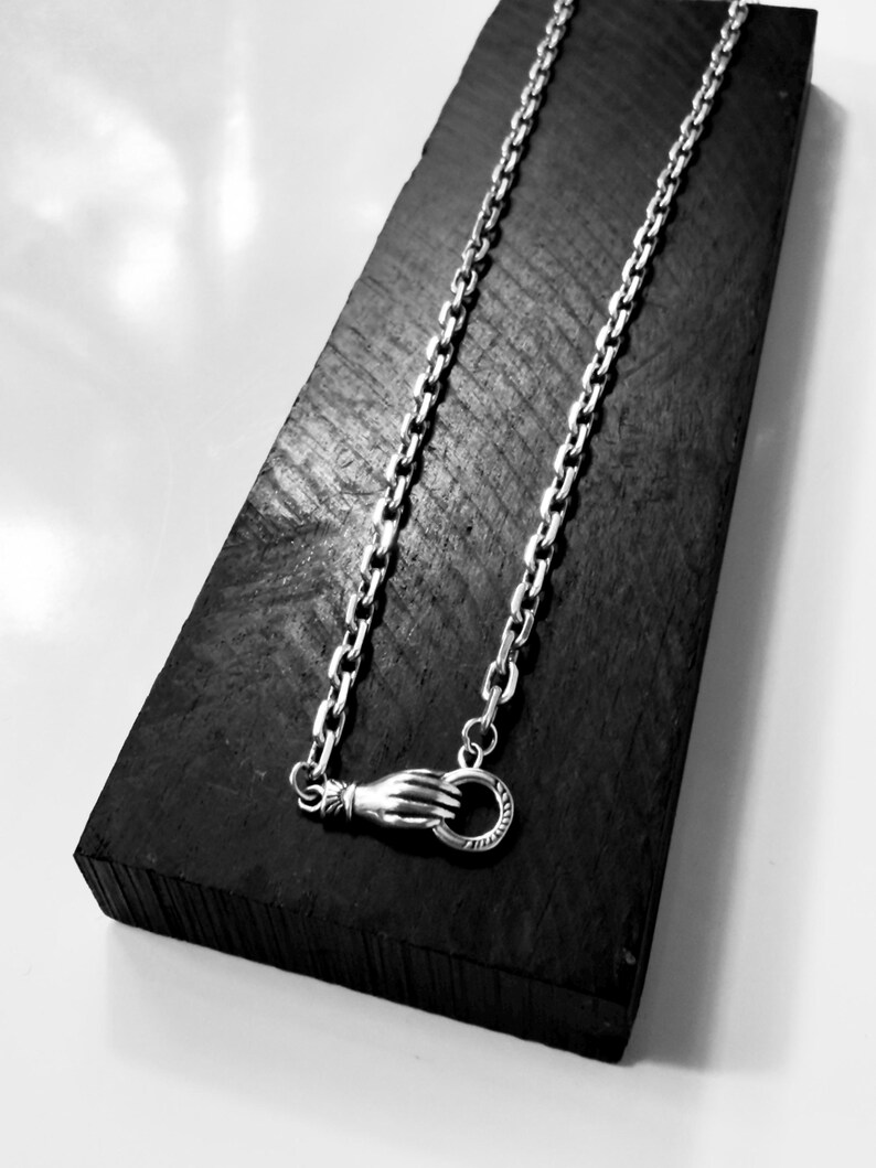 May include: A silver chain necklace featuring a clasp designed as a hand holding a circular ring. The metallic links are uniform and connected to the hand-shaped fastener, which secures the necklace with a circular loop mechanism.