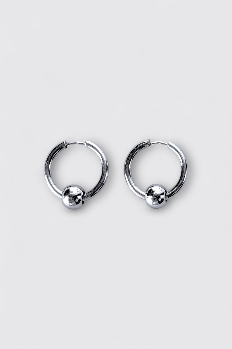May include: A pair of silver-toned hoop earrings, each featuring a smooth, polished spherical bead centered on the bottom curve of the circular hoop. The earrings have a reflective metallic finish and a simple, circular wire closure mechanism.